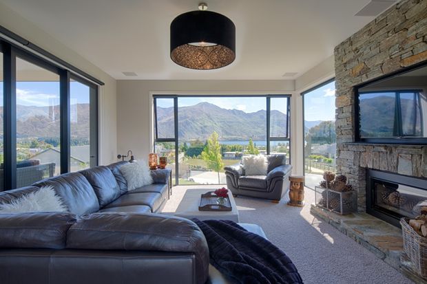 Bright, spacious living room with large floor-to-ceiling windows overlooking a mountainous lakeside landscape. The room features a large dark leather sectional sofa, matching armchair, carpeted flooring, a substantial rough-stone fireplace wall with integrated television, and a dark drum pendant light. The interior has a comfortable lodge-inspired character with strong natural views and abundant daylight.