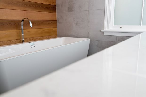 Close-up view of a contemporary bathroom featuring a freestanding white bathtub, chrome wall-mounted or deck-mounted faucet, large grey wall tiles, warm horizontal timber wall panelling, and a white foreground surface likely part of a vanity or benchtop. A frosted window with white trim provides natural light.