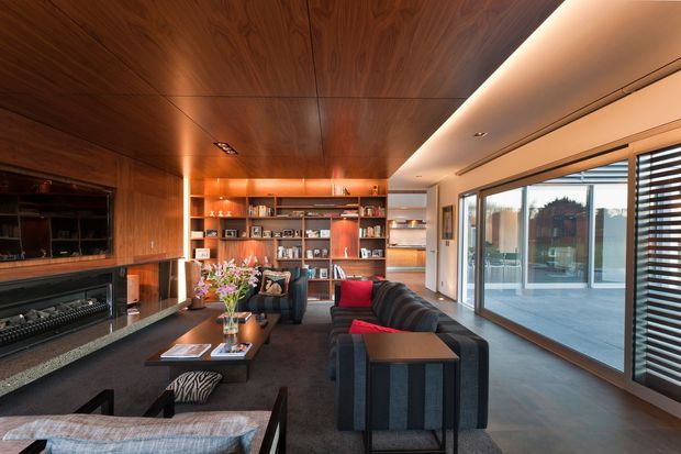 A refined contemporary living room with extensive dark timber wall and ceiling panelling, a built-in fireplace and television wall, full-height shelving and cabinetry, and a large charcoal sectional sofa. Warm concealed lighting highlights the timber joinery, while expansive glazed sliding doors and horizontal blinds open to a bright outdoor terrace. The room has a sophisticated, warm, media-focused atmosphere.