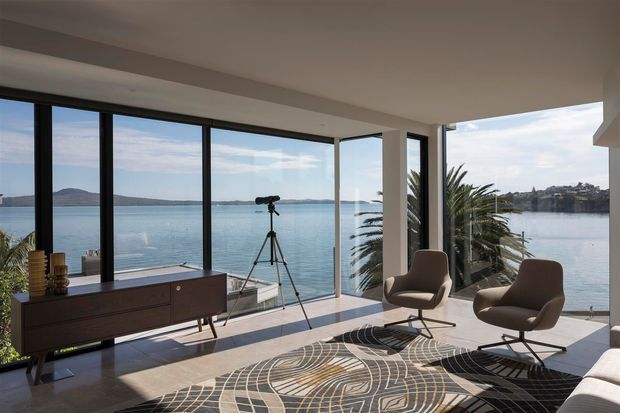 A bright, contemporary waterfront living room with expansive floor-to-ceiling corner glazing overlooking calm water and distant hills. The room contains a dark timber sideboard, two sculptural lounge chairs with ottomans, a patterned area rug, indoor palms, and a camera tripod near the windows. The space has a refined mid-century-inspired furnishing scheme, a light neutral interior, and strong indoor-outdoor visual connection.