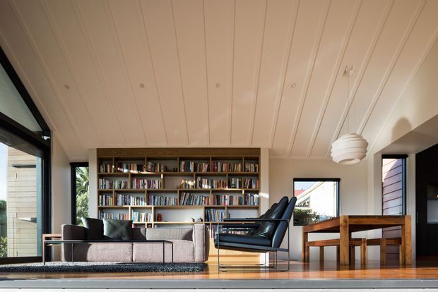 A bright, refined living room with a large built-in wall-to-wall bookcase, upholstered sofa, black leather lounge chair, timber dining table and chairs, and expansive black-framed glazing. The room features a warm timber floor, pale vertical-panel ceiling, neutral furnishings, and a sculptural pendant light. The open glazing provides views to the surrounding exterior.