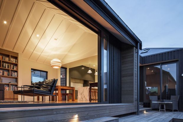 Twilight view of a contemporary residential extension opening onto a timber deck. The main space is an open-plan living and dining area with a built-in bookshelf, leather lounge chair, timber dining table, pendant lighting and a kitchen visible beyond. Large glazed openings connect the interior to the deck and a secondary dark-clad wing.