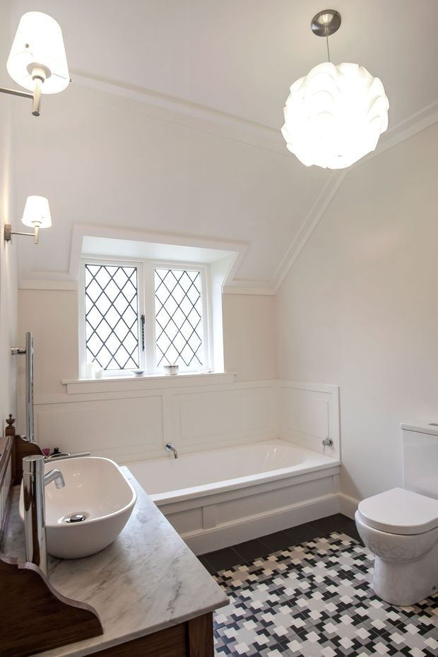 Bright, refined bathroom with a freestanding-style built-in white bathtub beneath leaded diamond-pane windows, a vessel sink on a marble-look benchtop with dark timber cabinetry, and a white toilet. White painted wall panelling and decorative ceiling cornices are paired with a bold geometric black, white and grey patterned floor tile. Contemporary pendant and wall lighting add sculptural detail.