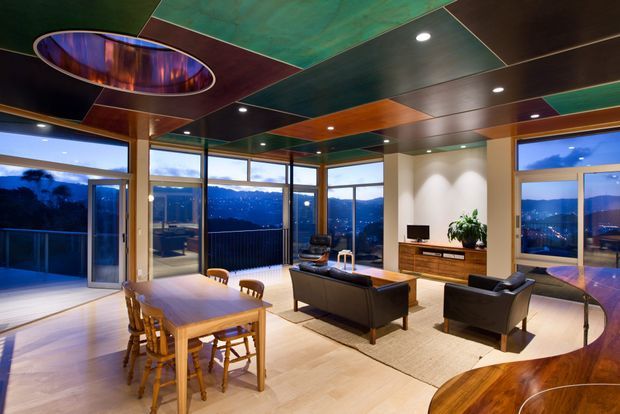 A dramatic open-plan living and dining area with expansive floor-to-ceiling glazing overlooking a mountainous landscape. The room features a timber dining table and chairs, dark leather seating, a timber media console, pale flooring, and a striking multicoloured geometric ceiling with recessed lighting and an oval illuminated feature.