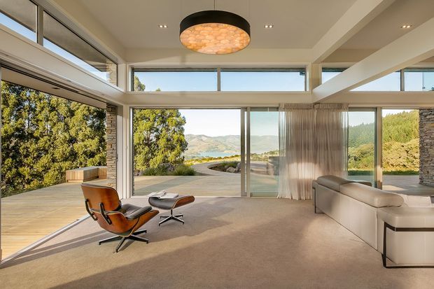 A bright, spacious contemporary living room with expansive floor-to-ceiling glazing and clerestory windows overlooking a landscaped hillside and water beyond. The room features a large neutral sectional sofa, a mid-century leather lounge chair with ottoman, broad carpeted flooring, a sculptural pendant light, exposed structural beams, and a partially visible stone-clad wall. Timber decking extends outside the glazing.