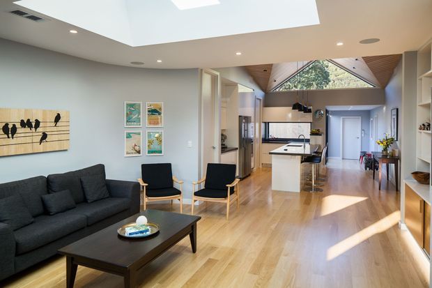 Open-plan contemporary home interior viewed from the living room, with a dark charcoal sofa and coffee table in the foreground, light timber flooring, pale walls, and a compact kitchen with a white island beyond. The space features a dramatic pitched timber-lined ceiling with triangular clerestory glazing, a large rectangular skylight, recessed lighting, built-in shelving, and a bright, connected living-kitchen layout.