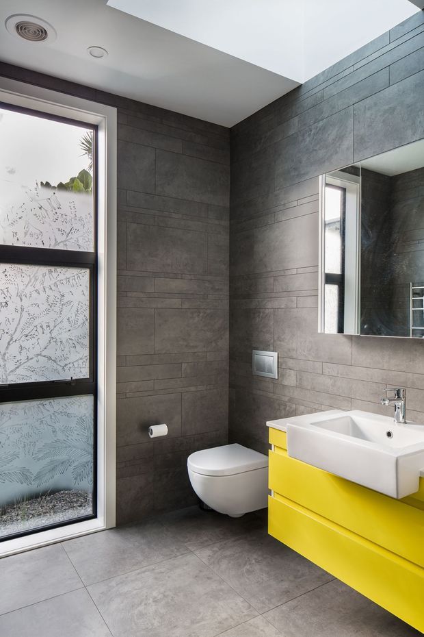 A contemporary bathroom with large-format dark grey tiled walls and floor, a wall-hung white toilet, and a bright yellow floating vanity with a white rectangular basin. A large mirror sits above the vanity, while a tall frosted patterned window provides natural light. The ceiling has recessed spotlights and an air vent.