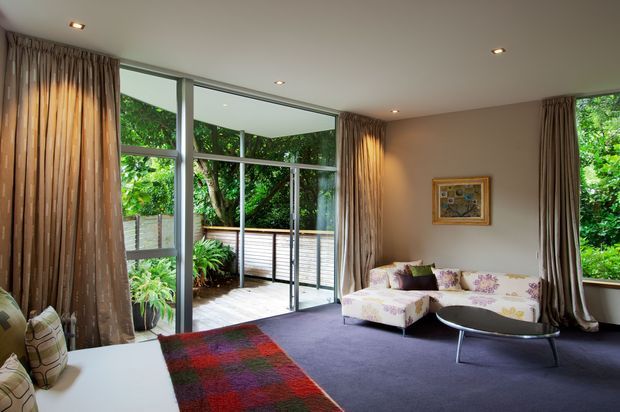 A spacious living room with floor-to-ceiling glazing and large sliding glass doors opening to a timber deck surrounded by dense greenery. The room features full-height taupe curtains, a deep purple carpet, a floral sectional sofa, a low oval coffee table, recessed ceiling lights, and a colourful patterned rug. The restrained modern shell is softened by patterned furnishings and warm natural views.