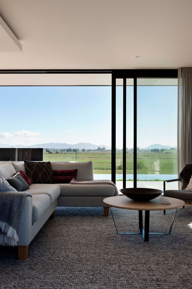 A contemporary living room with a large grey sectional sofa, dark accent cushions, a round timber coffee table with a black sculptural base, and a textured neutral rug. Full-height black-framed glazing opens to expansive green rural fields, distant mountains, and a bright blue sky. The room has a restrained neutral palette, clean lines, and abundant natural light.
