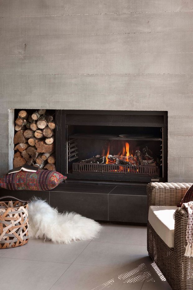 A cozy contemporary living room centered on a built-in black fireplace with a visible fire, set into a textured pale grey wall. An integrated open firewood niche sits beside it, while dark tiled hearth surfaces, a patterned cushion, woven rattan seating, and a sheepskin rug add rustic and bohemian warmth.