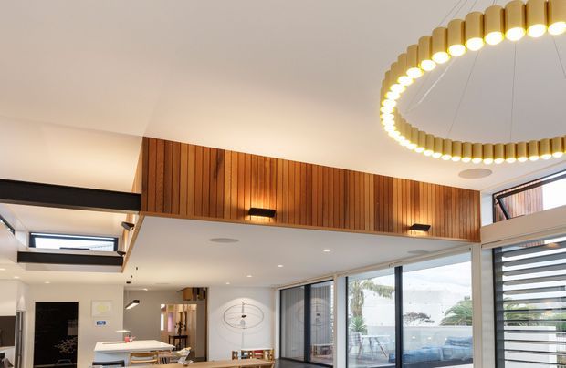 A bright contemporary open-plan living and dining space with a high white ceiling, extensive floor-to-ceiling glazing, and a warm vertical timber feature wall wrapping the upper level. A large circular pendant light with repeated gold-toned elements hangs prominently overhead. The room includes a dining table, kitchen island, built-in cabinetry, and a glazed connection to an outdoor terrace or pool area. Horizontal louvres and clerestory-style windows add architectural detail.