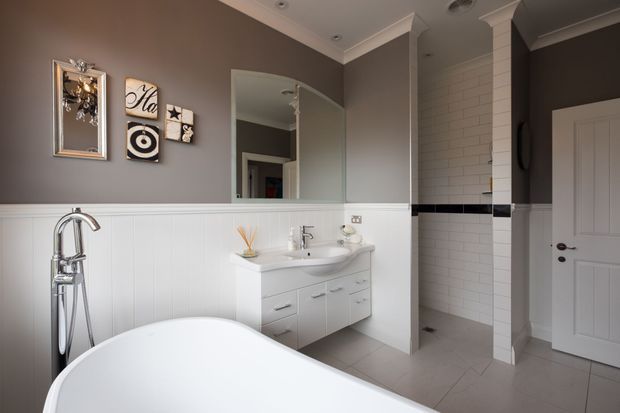 A refined bathroom with a freestanding white bathtub in the foreground, a chrome floor-mounted bath filler, a floating white vanity with integrated basin, and a large wall mirror. White vertical wall panelling is paired with taupe-grey painted walls, while the shower area features white subway tiles with a horizontal black accent strip. A white paneled door and pale tiled floor complete the space.