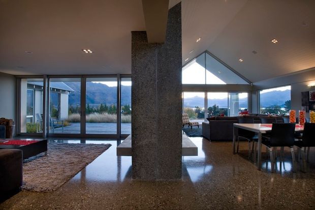 Spacious open-plan contemporary interior with polished terrazzo flooring, a large central stone-clad structural column and integrated bench, expansive floor-to-ceiling glazing, and a dramatic triangular clerestory window. The room combines living and dining zones and opens toward a landscaped exterior with mountain views. Recessed lighting, dark upholstered furniture, and warm neutral finishes create a refined, modern atmosphere.