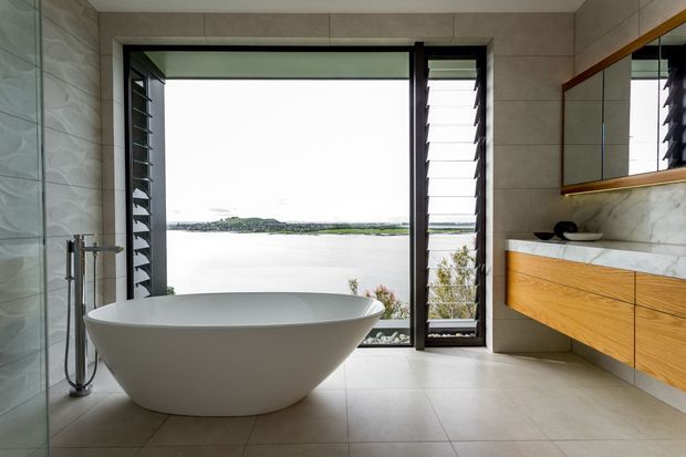 Contemporary bathroom with a freestanding white bathtub positioned beside full-height louvred glazing overlooking a body of water. The room features large light-grey stone-look wall and floor tiles, a timber floating vanity with a pale stone benchtop, a frameless glass shower enclosure, and a large mirrored cabinet. The space is bright, refined, and strongly connected to the waterfront outlook.