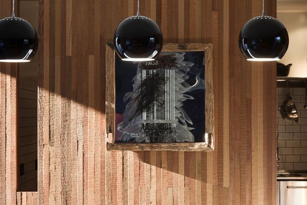 Interior view featuring a richly textured vertical timber feature wall with varied warm wood tones and a framed monochrome artwork. Three glossy black globe pendant lights hang across the foreground. Partial views of a dark-tiled adjacent area and kitchen joinery appear at the sides.