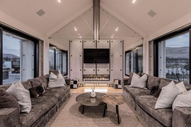A spacious, symmetrical contemporary living room with paired grey upholstered sofas facing a central television and fireplace joinery. Full-height black-framed glazing on both sides provides expansive views of a rugged natural landscape. The room has a vaulted white ceiling with exposed central ductwork, recessed lighting, pale built-in wall panels, a large neutral rug, and nested dark coffee tables.