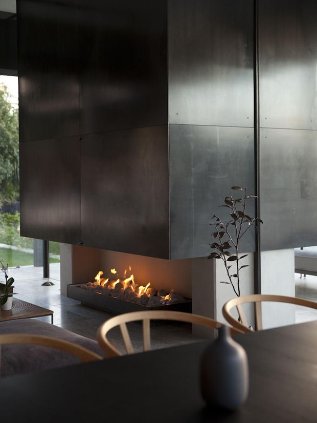 A dark, refined contemporary interior centered on a linear open fireplace beneath a large suspended dark metal-clad volume. The room includes floor-to-ceiling glazing, a dark dining table, curved timber dining chairs, a simple ceramic vase, and views to greenery outside. The fireplace and reflective metal surfaces create a warm focal point within an otherwise moody, minimalist space.