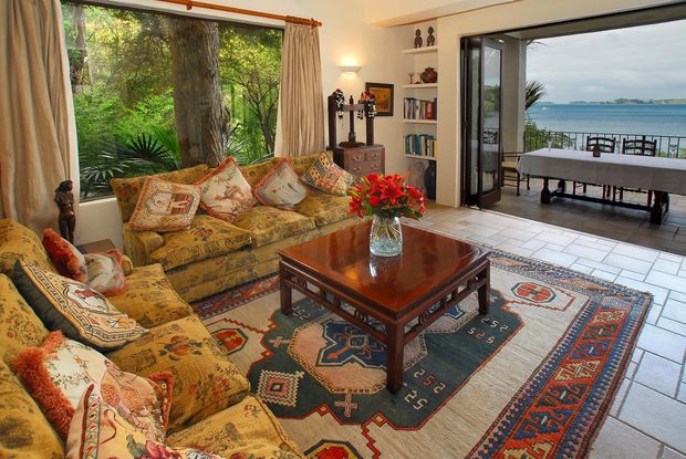 A richly furnished living room with patterned upholstered sofas, an ornate dark wood coffee table, layered geometric rugs, and decorative shelving. Large windows overlook lush vegetation, while open folding doors connect to a tiled outdoor terrace with dining furniture and a broad coastal water view.