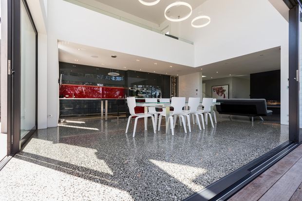 Bright, expansive contemporary open-plan interior viewed through fully opened glazed doors. The space combines a dining area with white chairs and table, a sleek kitchen with a bold red island or splashback, a dark lounge sofa, polished terrazzo flooring, a double-height void with upper-level glazing, and sculptural circular pendant lights.