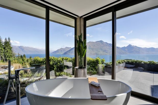 A freestanding white bathtub occupies the foreground of a bright contemporary bathroom, positioned beside expansive black-framed corner glazing with panoramic views of a blue lake and surrounding mountains. A dark timber bath caddy and tall indoor plant add warmth, while a paved terrace and planted screening are visible outside.
