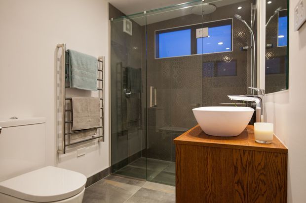 Compact contemporary bathroom featuring a frameless glass walk-in shower, dark patterned feature tiles, grey floor tiles, a wall-mounted heated towel rail, white toilet, and a freestanding white vessel basin on a warm timber vanity. Recessed ceiling lighting and small high windows provide a clean, refined atmosphere.