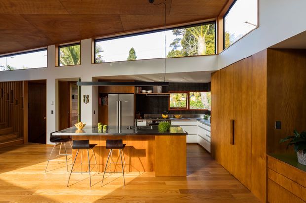 Warm, contemporary open-plan kitchen with a timber-clad ceiling, extensive high-level glazing, rich timber flooring, and matching timber cabinetry. A central island with dark stone benchtop and bar seating faces stainless-steel appliances and white cabinetry, while a timber-lined wall provides additional integrated storage. The space is bright and connected to greenery outside.