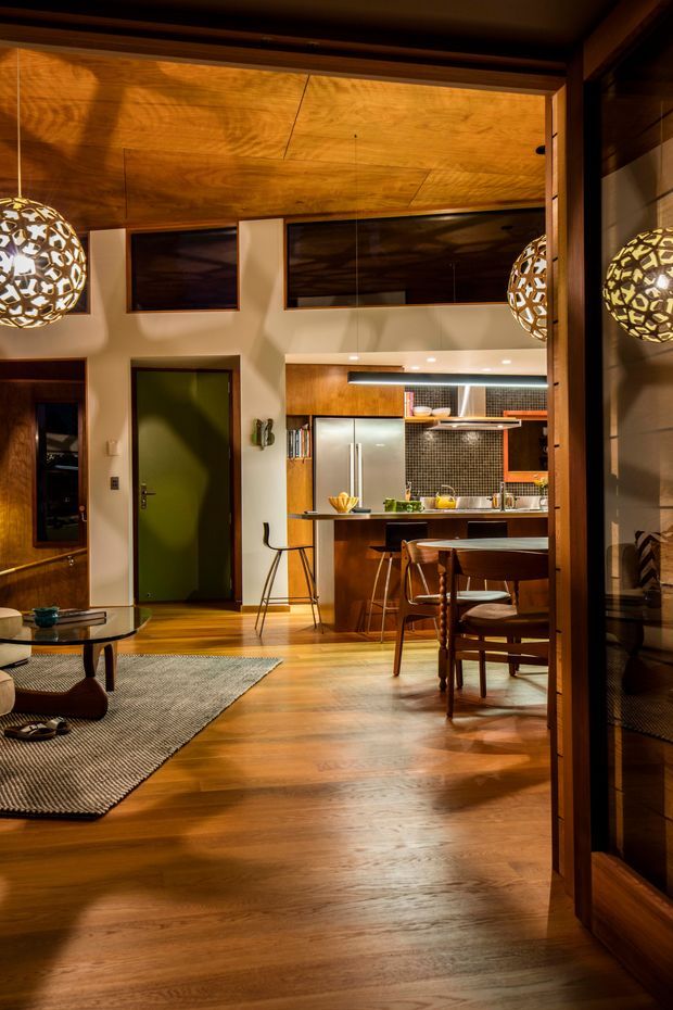 Warm, richly detailed open-plan interior combining a living room, dining area, and compact kitchen. The space features extensive timber flooring and ceiling panelling, dark timber-framed glazing and joinery, a green internal door, a stainless-steel refrigerator, a dark mosaic-tile splashback, timber kitchen cabinetry, a peninsula with bar seating, mid-century-style timber dining furniture, and sculptural pendant lights. The view is photographed from within the home through a timber-framed opening, with a mirror or reflective glazing visible at right.