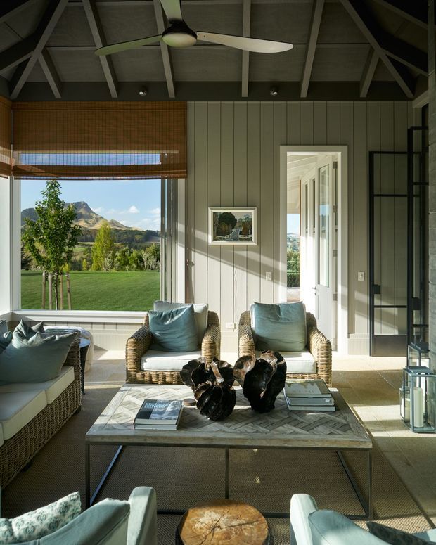 A bright, relaxed living room with large windows overlooking a landscaped lawn and mountainous scenery. Wicker armchairs and sofa seating surround a substantial marble-topped coffee table, while vertical painted wall panelling, exposed roof rafters, a ceiling fan, and a glass-panel door create a refined indoor-outdoor feel.