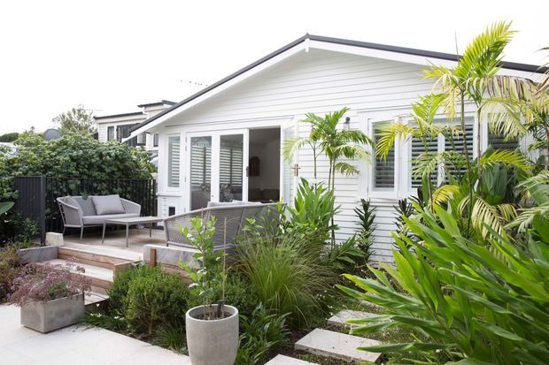 Single-storey white weatherboard house with a pitched gable roof, broad glazed doors and windows, and a small timber deck furnished with outdoor seating. Lush tropical and subtropical planting surrounds the deck, with stepping stones leading through the garden.