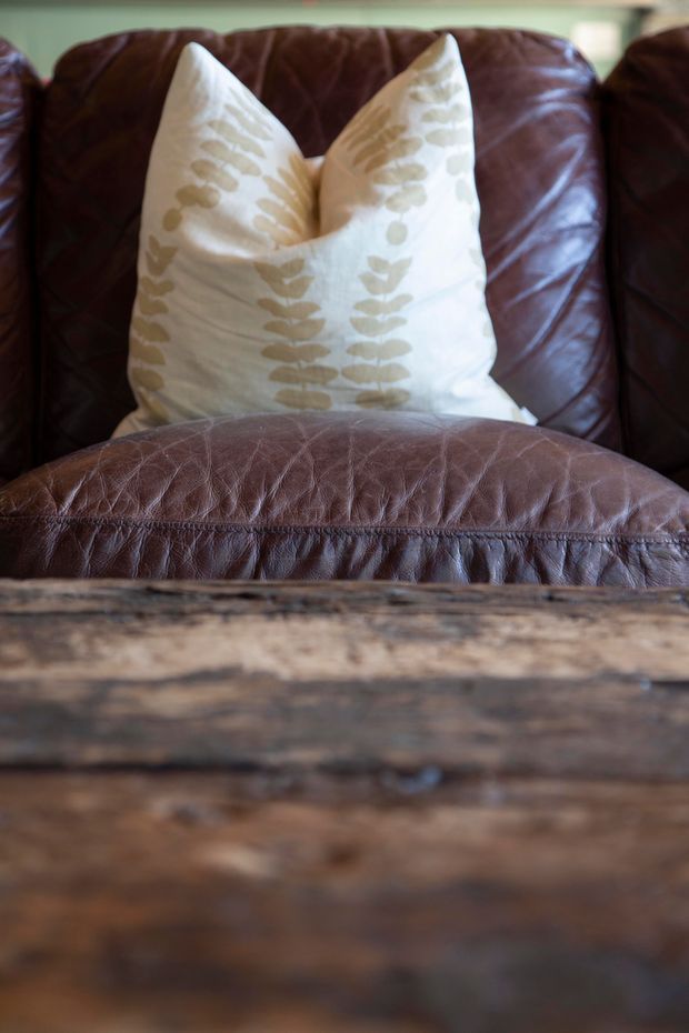 Close-up interior detail of a deep brown leather armchair with a cream decorative cushion featuring a muted botanical pattern. A rustic, heavily grained dark timber table occupies the foreground, creating a warm, tactile composition.