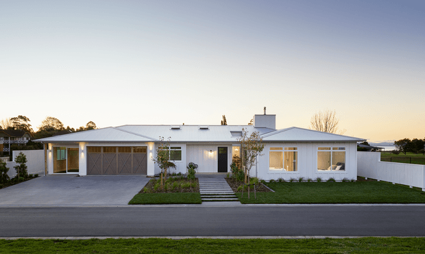 Single-storey white residential house viewed from across the street at dusk. The home has a low-pitched hipped metal roof, white rendered walls, broad rectangular windows, a covered entry, attached double garage with timber-look doors, a central landscaped path and neatly maintained lawns. A white boundary fence and open rural or suburban surroundings are visible in the background.