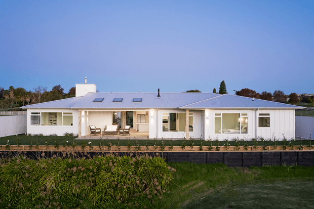 Single-storey contemporary residential home viewed from the rear, with a broad low-pitched metal roof, white exterior walls, extensive floor-to-ceiling glazing and sliding doors opening onto a long timber deck. Multiple roof skylights and a central raised roof element are visible. Landscaped planting and lawn sit in the foreground.