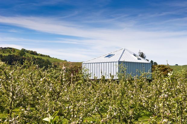 A small, modern greenhouse-like structure with a steep pyramidal metal roof and glazed or translucent vertical wall panels, set amid a flowering orchard in rolling green countryside. The bright outdoor scene emphasizes the crisp geometric building form against the landscape and blue sky.