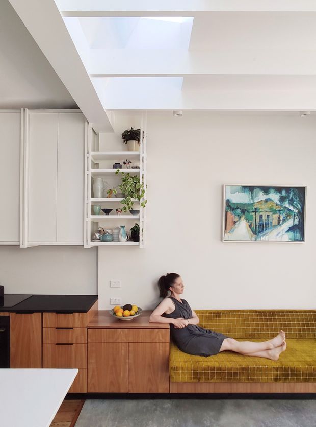 Bright, compact open-plan living space with a built-in timber kitchen and cabinetry, white upper cabinets, open display shelving with plants and ceramics, and a mustard-yellow built-in banquette. A woman is seated on the banquette beneath a colourful framed artwork. White walls and ceiling beams surround a large skylight, creating an airy modern interior.