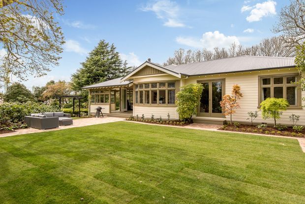 A single-storey suburban home viewed from a landscaped lawn, with cream horizontal weatherboard cladding, a low-pitched metal roof, projecting gabled bay, and extensive timber-framed windows. A paved patio with outdoor seating sits to the left, while established garden planting surrounds the house.