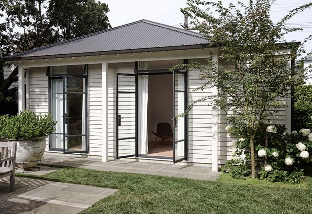 Single-storey white weatherboard building with a low-pitched dark metal hipped roof, broad eaves, and tall black-framed glazed doors opening onto a paved garden terrace. The exterior is viewed from a landscaped outdoor area, with planting, hydrangeas, lawn, and a small outdoor seating area visible. The open doors reveal a simple interior room beyond.