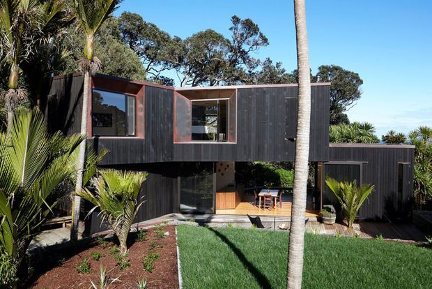 Exterior view of a contemporary two-storey home set within lush subtropical landscaping. The building has dark vertical timber cladding, warm reddish timber window and door frames, projecting and recessed volumes, and a small timber deck at the lower level. Palm trees, clipped lawn and garden beds surround the house.