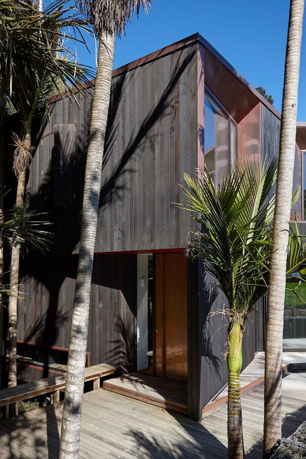 Exterior view of a compact contemporary multi-level residence clad in dark vertical timber boards, with angular roof forms, large glazed openings, and a timber deck surrounded by palm vegetation. Strong sunlight creates pronounced shadows across the facade and decking.