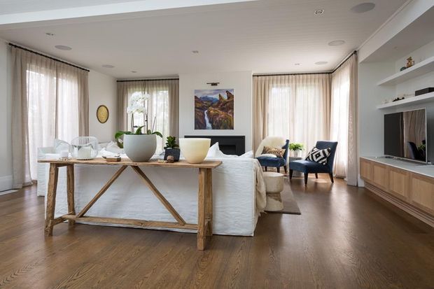Bright, refined open-plan living room with warm timber flooring, white upholstered seating, a rustic timber console table behind the sofa, floor-to-ceiling sheer curtains, built-in white shelving and timber media cabinetry. A fireplace, artwork, armchairs and decorative plants complete the relaxed contemporary interior.