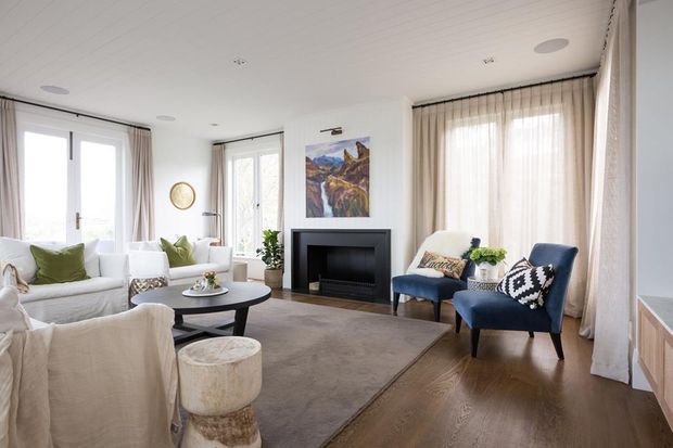 Bright, refined living room with white upholstered seating, pale green cushions, navy accent armchairs, a round black coffee table, a contemporary black fireplace surround, full-height curtains, timber flooring, and a large neutral rug. The room includes generous glazing and recessed ceiling lighting, with a colourful landscape artwork above the fireplace.