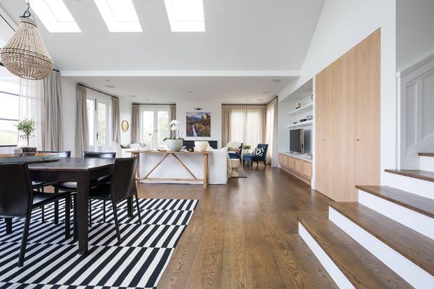 Bright open-plan residential interior combining a dining area and living room, with white walls, warm timber flooring, a black-and-white geometric rug, built-in timber cabinetry, skylights, and a staircase leading upward. Large windows and light curtains create an airy atmosphere, while a woven pendant light adds natural texture.