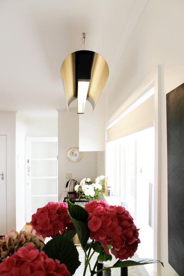 Bright white interior with a compact kitchen or kitchenette visible in the background, large windows with roller blinds, and a striking suspended sculptural pendant light with a gold-toned interior. Foreground hydrangea flowers partially obscure the lower portion of the room. White built-in shelving, pale cabinetry, and a small round wall clock contribute to the clean, light-filled setting.