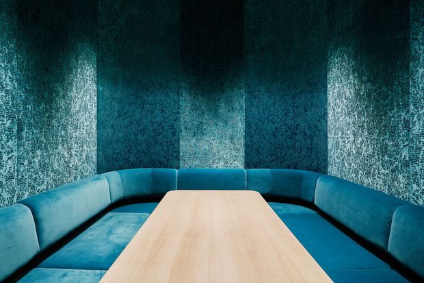 A compact, enclosed lounge or meeting nook with a large U-shaped upholstered banquette in vivid teal-blue, surrounding a pale timber rectangular table. The walls and ceiling are covered in dark, heavily textured blue-green material, creating an immersive, cocoon-like atmosphere with dramatic contrast between the dark enclosure, bright seating, and light wood tabletop.