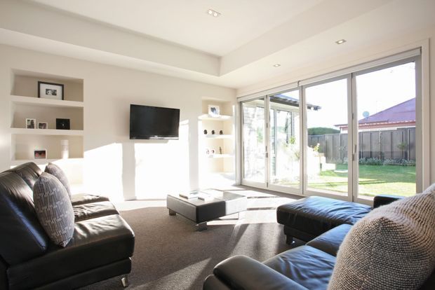 Bright contemporary living room with a large black leather sectional sofa, low square coffee table, wall-mounted television, recessed display shelving, and expansive sliding glass doors opening to a sunny backyard. The room has light neutral walls, carpet flooring, and abundant natural light.
