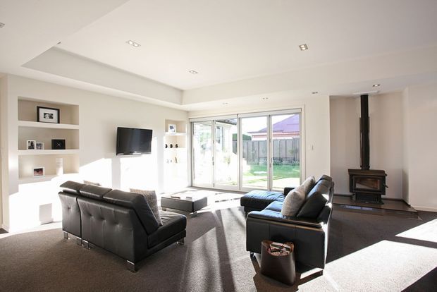 Bright contemporary living room with two black leather sofas, a wall-mounted television, built-in white display shelving, recessed ceiling lights, and large sliding glass doors opening to a garden. A freestanding wood-burning stove with a dark flue forms a feature on the right, while dark carpet and neutral walls create a clean, spacious interior.