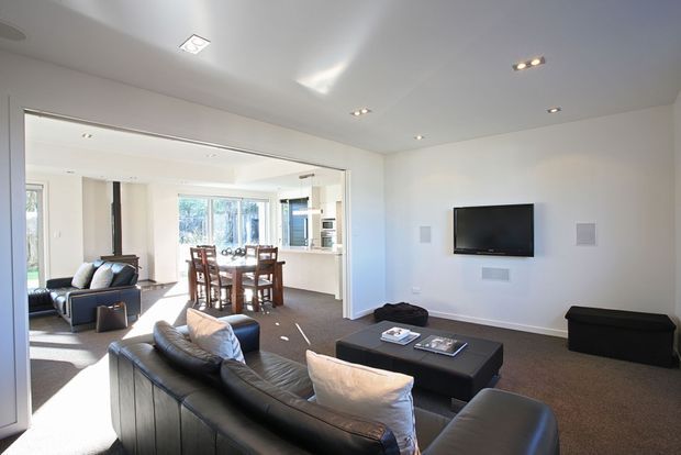 Bright open-plan living and dining area with a contemporary minimalist fit-out. The living room has black leather sofas, a large black ottoman coffee table, wall-mounted television and concealed speakers, opening directly to a dining area with a timber dining table and chairs. White walls and ceiling, recessed downlights, broad internal opening and dark carpet create a clean, modern interior.
