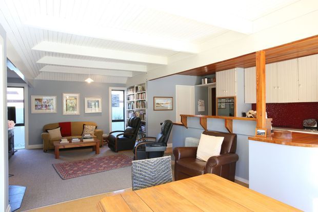 Open-plan residential interior combining a living room, dining area and kitchen. The room features a white timber-lined ceiling with exposed beams, grey painted walls, timber-framed cabinetry and counters, a dark red mosaic splashback, carpeted living area, and a warm timber dining table. The furnishings include a sofa, leather armchairs, coffee table, patterned rug, and built-in shelving.