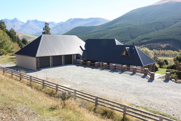A dark-roofed, single-storey rural building complex set in a broad gravel forecourt, with mountain ranges and steep forested hills in the background. The main structure has a steep pitched roof, dark cladding, a light metal-roofed wing, covered outdoor verandah, and timber fencing in the foreground.