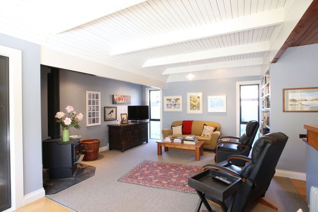 Bright, inviting living room with a white-painted timber-lined ceiling featuring exposed beams and recessed skylight-like panels. The room has grey walls, carpet flooring, a dark timber entertainment cabinet, a tan sofa with colourful cushions, two black leather armchairs, a timber coffee table, patterned rug, framed artwork, and large windows providing natural light.