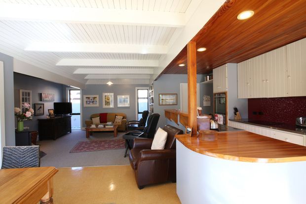 Open-plan mid-century-inspired interior combining a living room and kitchen. The space features a white timber-lined ceiling with exposed beam forms, warm timber cabinetry and ceiling lining, a curved white kitchen island with timber benchtop, dark red textured splashback, grey walls, polished concrete or terrazzo-like flooring, and a furnished lounge area with leather armchairs and a sofa.